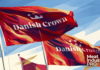 Sokołów полностью интегрирован в группу Danish Crown Sokołów полностью интегрирован в группу Danish Crown — живописная иллюстрация: три развевающихся красных флага с белой эмблемой короны и надписью «Danish Crown» на фоне голубого неба с широкими белыми, бирюзовыми и золотистыми диагональными плоскостями; полотнища переданы плотными мазками алого, бордового, терракотового и кремового цвета на металлических флагштоках; логотип Meat Industry PRO белым шрифтом в правом нижнем углу